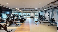 Located in Baltimore, MD, this fitness center offers a thoughtfully designed, single-level space outfitted for a full range of workout routines. The open floor plan is finished with a combination of warm hardwood flooring and durable rubber matting, creating distinct zones for cardio, strength training, and functional exercise.

The cardio area is lined with treadmills, elliptical machines, and stationary spin bikes, all positioned to take advantage of large windows that flood the space with natural light and views of the surrounding urban environment. Full-length mirrors along the walls enhance the sense of openness and allow users to monitor their form throughout their workout.

The strength and free weights section features a tiered dumbbell rack, adjustable benches, a cable machine, a squat rack with weight plates, and select resistance machines including a leg extension/curl unit — providing comprehensive options for both beginners and experienced athletes.

Exposed ductwork runs along the ceiling, lending the space an industrial aesthetic that is softened by recessed lighting, track lighting, and clean white walls. A built-in water station with a bottle filler is conveniently located near the entrance, alongside storage shelving. Steel-framed glass doors provide a clean, modern entry point and connect the fitness center to the broader facility corridor.