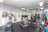 This fitness center is a dedicated, single-floor workout facility designed to support a well-rounded exercise routine. The space is characterized by its bold, energetic aesthetic — walls are painted in a light gray tone and accented with a distinctive horizontal orange and gray stripe that runs throughout the room, lending a dynamic, motivating atmosphere.

The facility is outfitted with a solid selection of cardio equipment, including treadmills, an elliptical trainer, and a stair climber, all arranged to maximize floor space and ease of movement. A multi-station cable strength machine anchors one side of the room alongside medicine balls and additional accessories, providing a comprehensive strength-training option. Full-length mirrors line the walls, enhancing the sense of openness and allowing users to monitor their form during workouts.

Natural light filters in through multiple sets of white-framed French doors and windows, keeping the environment bright and inviting. One set of French doors opens directly to an outdoor pool area, offering convenient access and pleasant views of greenery beyond. A wall-mounted flat-screen television adds an entertainment element for users during cardio sessions.

Crown molding along the ceiling and dark rubberized flooring complete the space, balancing practical gym functionality with a polished, well-maintained appearance. Fitness center rules are posted on the wall for easy reference.