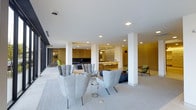 This single-floor space presents a refined contemporary design characterized by clean lines, an open-plan layout, and a carefully curated aesthetic. The interior is anchored by a light-toned floor that stretches across the entire level, lending an airy, expansive quality to the environment. Structural white columns define zones within the open floor plan without interrupting the sense of flow between areas.

The main lounge area is furnished with elegant, high-backed upholstered armchairs in soft grey tones, arranged to encourage both comfort and conversation. A dark navy sofa anchors a secondary seating grouping, complemented by additional accent chairs in green and neutral tones that introduce subtle pops of color throughout the space.

A prominent feature is the warm wood-paneled wall that lines the back portion of the interior, providing a natural contrast to the otherwise white and neutral palette. This section also incorporates what appears to be a reception or bar counter with illuminated detailing, suggesting a hospitality or lounge function. Recessed ceiling lighting is distributed evenly throughout, ensuring consistent, well-balanced illumination across all areas.

Large openings along the perimeter hint at connections to exterior spaces, allowing natural light to filter in and enhance the overall brightness of the interior. The result is a polished, modern environment with a welcoming yet sophisticated ambiance.