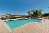 Located at 12730 W Camelback Rd in Litchfield Park, Arizona, this single-level outdoor community amenity area offers a resort-style experience designed for relaxation and recreation under the sunny Arizona sky. At the heart of the space is a generously sized swimming pool with a freeform shape, finished with striking blue tile accents and a wide, paver-tiled deck ideal for lounging. An attached circular spa sits adjacent to the main pool, providing a dedicated space for relaxation. Tall, mature date palms and weeping trees frame the area, casting natural shade and lending a lush, desert-oasis ambiance. A covered ramada with a standing-seam metal roof serves as the central gathering hub, offering shaded seating and dining areas for groups. Nearby, a built-in outdoor kitchen with a gas grill provides convenient amenities for outdoor entertaining. Comfortable lounge chairs are arranged throughout the sun deck, accommodating multiple guests at once. A dedicated restroom facility with Spanish-style tile roofing is conveniently located poolside, accompanied by a drinking fountain. Additional recreational touches include a cornhole game set, adding a casual, social dimension to the space. The surrounding landscape features manicured lawn panels, desert plantings, and perimeter fencing, creating a private and well-appointed outdoor retreat.