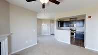 This single-floor residence offers a thoughtfully designed open-concept layout that connects the living and kitchen areas in a cohesive, functional flow. The living area is finished with plush neutral carpeting and soft beige walls, creating a warm and inviting atmosphere. A ceiling fan with a light fixture anchors the main living space, providing both comfort and illumination. An elegant chandelier marks the transition toward the entryway, which features a clean, classic door framed by crisp white trim.

The kitchen is positioned on an elevated platform with a raised breakfast bar that defines the space without compromising the open feel. Dark espresso cabinetry provides a striking contrast against the light countertops, while stainless steel appliances — including a gas range and over-the-range microwave — add a modern, polished touch. The kitchen area is finished with hardwood-style flooring, distinguishing it from the carpeted living zone and reinforcing the intentional design of the space.

Throughout the home, neutral tones and clean lines work together to create a bright, understated aesthetic that is both versatile and welcoming. The overall layout is efficient yet spacious, making excellent use of the single-level floor plan.