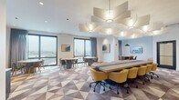 Located in Chicago's Uptown neighborhood at 4613 N Broadway St, this contemporary professional floor offers a thoughtfully designed suite of collaborative and private workspaces. The main open area serves as the centerpiece, featuring a long communal conference table lined with mustard-yellow upholstered chairs beneath a striking sculptural ceiling fixture composed of layered, geometric light panels. The geometric-patterned carpet in muted tones of gray, mauve, and cream adds visual interest throughout the floor. Floor-to-ceiling windows along the perimeter flood the space with natural light and frame expansive views of the Chicago skyline and surrounding neighborhoods. Multiple round tables with gray-upholstered chairs are arranged near the windows, providing flexible seating for group discussions or informal gatherings. A lounge area features a channeled gray sectional sofa paired with sleek black pedestal tables, positioned beside a mirrored partition wall with a textured metallic accent surface. Private focus rooms with glass-panel doors and smart room-booking panels offer individual workstations for concentrated work. A dedicated conference room with a round wooden table, warm leather chairs, an oversized pendant light, and a wall-mounted display screen provides an ideal setting for smaller meetings. The overall ambiance is polished, bright, and professional.