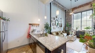 Located at 1101 Hawthorne Lane in Charlotte's vibrant Plaza Midwood neighborhood, this one-bedroom, one-bathroom loft delivers a compelling blend of industrial heritage and eclectic character. Soaring ceilings amplify the sense of openness throughout, while dramatic floor-to-ceiling, steel-framed windows bathe every room in abundant natural light and offer lush green views outdoors.

The living area showcases exposed brick walls and structural columns that speak to the building's historic roots, creating a warm, textured backdrop for the open-concept layout. A gallery-style wall arrangement adds artistic personality to the space.

The kitchen is well-appointed with dark cabinetry, granite countertops, stainless steel appliances, and a peninsula with bar seating — ideal for both casual dining and meal preparation. Pendant lighting highlights the generous counter space and galley-style layout.

The bedroom continues the loft's industrial aesthetic, featuring the same soaring ceilings, exposed brick, and oversized windows that invite natural light and outdoor scenery. The room comfortably accommodates a full sleeping and work area.

The bathroom is finished with a bold, classic checkerboard tile floor, a shower/tub combination, and clean white walls. Throughout the loft, mixed flooring materials and high ceilings reinforce the distinctive, character-rich atmosphere of this unique Charlotte residence.