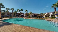 Located at 13444 Gran Bay Pkwy in Jacksonville, Florida, this outdoor amenity space offers a well-appointed resort-style experience designed for relaxation and recreation. At its centerpiece is a generously sized swimming pool with crystal-clear turquoise water, surrounded by a broad paver deck lined with lounge chairs and market umbrellas for shade. Tall palm trees are thoughtfully interspersed throughout the pool area, lending a tropical ambiance beneath the abundant Florida sunshine.

Adjacent to the pool, a covered outdoor lounge area features a coffered ceiling with a ceiling fan, providing a comfortable retreat from the sun. This shaded space is furnished with stylish seating—including sofas and accent chairs in coastal-inspired blue and aqua tones—arranged around a coffee table, ideal for socializing or unwinding. French doors connect this covered patio to interior facilities.

Additional paver patios extend from the building, finished with stacked stone exterior accents and offering open-air seating beneath umbrellas. The surrounding landscaping features carefully maintained plantings, decorative gravel beds, and large potted arrangements that enhance the visual appeal of the grounds. The overall design creates a cohesive, upscale outdoor environment suited to the warm Jacksonville climate.