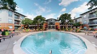 Located in Dallas, this outdoor amenity space centers around a generously sized resort-style pool that serves as the focal point of a thoughtfully designed courtyard environment. The pool area is defined by a broad, stamped concrete sun deck outfitted with vibrant orange chaise lounges and colorful seating clusters, creating a lively yet relaxed atmosphere ideal for outdoor living.

Mature crape myrtles and tall shade trees are strategically placed throughout the courtyard, providing natural canopy and greenery that soften the surrounding multi-story brick and siding residential buildings. Colorful flowering plants and well-maintained landscaping beds add seasonal interest along the pool perimeter and walkways.

A standout architectural feature is the clubhouse building, which anchors the courtyard with a dramatic curved roofline and soaring floor-to-ceiling glass facade that floods the interior with natural light and creates a strong visual connection between indoor and outdoor spaces. A covered pergola walkway runs alongside the clubhouse, offering a shaded path between the building and the pool deck. Patio dining areas with market umbrellas in red provide additional outdoor social space. The overall ambiance is that of a well-appointed, resort-inspired community retreat designed for year-round enjoyment in the Dallas climate.