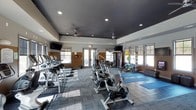This fitness center offers a comprehensive workout environment designed to support a wide range of exercise routines. The space features a dark, painted ceiling with recessed lighting and ceiling fans, creating a focused yet comfortable atmosphere. Large windows line the walls, allowing natural light to fill the room and providing a bright, energizing setting for workouts.

The cardio zone is prominently featured along one side of the room, lined with a row of treadmills ready for endurance training. A cluster of stationary cycling bikes occupies the central floor space, ideal for spin workouts and low-impact cardio sessions. Toward the rear of the facility, a multi-station strength training machine is available for resistance and weight training exercises.

A dedicated stretching and floor exercise area is set up along one wall, complete with yoga mats and stability balls, providing space for flexibility training, core work, and cool-down routines. Wall-mounted flat-screen televisions are positioned throughout the space, allowing members to stay entertained during their workouts. The durable, interlocking tile flooring throughout the facility is both functional and easy to maintain, while the overall layout ensures ample room to move freely between equipment stations.