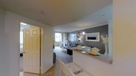 This single-floor residence offers a thoughtfully designed open-concept layout that connects the key living areas into a cohesive, flowing space. A breakfast bar serves as a natural divider between the kitchen and the dining area, providing both functional seating and a sense of definition without sacrificing openness. The dining space features stylish white chairs and elegant table settings, making it ideal for casual meals or entertaining guests.

The living room is anchored by a comfortable sofa and accented by a bold charcoal feature wall adorned with a framed mirror and contemporary artwork, adding depth and visual interest to the space. Warm pendant lighting and a combination of ambient light sources create an inviting atmosphere throughout. A large window and sliding glass door bathe the living area in natural light and provide direct access to an outdoor balcony, extending the living space beyond the interior.

Soft carpeting in the living area adds warmth and comfort underfoot, while the overall neutral color palette — highlighted by crisp whites and cool grays — lends the space a clean, modern aesthetic. The entry hallway offers a welcoming transition into the home, with a full door providing privacy and separation from the main living spaces. Throughout, the design balances style and practicality, making this residence both visually appealing and highly functional.