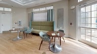 This single-floor space is thoughtfully designed to serve as a comfortable communal environment, combining several distinct functional areas beneath one roof. Light wood-look flooring flows consistently throughout, lending warmth and visual continuity to the entire layout.

The lounge area features a tall, high-backed green sofa paired with teal patterned accent chairs and a round tulip-style table, creating an inviting social setting. Decorative embossed wall panels and soft drapery in muted teal add refined detail. An entry foyer with panel-style doors, transom windows above, and a mounted AED unit offers a welcoming and safety-conscious arrival point.

A dedicated media nook is furnished with a low media console, dual monitors, and sleek white swivel chairs, providing a quiet spot for screen-based activities. The kitchen area is well-equipped with white shaker cabinetry, dark countertops, stainless steel appliances including a French door refrigerator and dishwasher, and a large window that fills the space with natural light.

A dining area with a pedestal table and upholstered chairs sits adjacent to French doors that open directly to an outdoor patio, offering views of greenery and outdoor seating. Throughout, the space balances practicality with a cohesive, contemporary aesthetic.