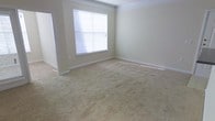 This single-floor space presents a clean, understated interior defined by neutral design choices and classic architectural details. The main room is finished with wall-to-wall beige carpeting that lends a warm, soft feel underfoot. Smooth, light-colored walls are accented by white crown molding along the ceiling line, adding a touch of traditional elegance to the otherwise minimalist space. A centrally positioned window fitted with horizontal blinds allows natural light to filter in, casting soft illumination across the room. White baseboards run along the perimeter, complementing the overall neutral color palette. The room is generously proportioned, offering a versatile, open layout well-suited to a variety of furniture arrangements. A doorway is visible along one wall, suggesting straightforward connectivity to adjacent areas of the home. The overall ambiance is quiet and neutral, providing a blank canvas that can be easily personalized. The combination of crown molding, clean lines, and soft carpeting gives the space a comfortable, move-in-ready character suited to residential living.