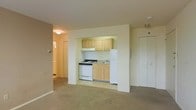 This single-floor residential unit offers a practical and functional layout designed to maximize the use of available space. The main living area is finished with wall-to-wall carpet and neutral-toned walls that lend a calm, understated ambiance throughout.

The kitchen is neatly tucked into a recessed nook, creating a defined cooking area without enclosing it entirely from the living space. It features light wood upper and lower cabinetry providing ample storage, a white gas range, a single-basin sink, and a full-size white refrigerator. The kitchen floor transitions to a smooth tile surface, distinguishing it from the carpeted living area.

A hallway visible from the main living area leads to additional rooms, suggesting a separation between living and private spaces. A flush-mount ceiling light provides overhead illumination in the living area, while the overall layout benefits from a straightforward, easy-to-navigate floor plan.

A door located on the far wall of the living area, likely a closet or utility space, adds to the storage options available within the unit. The clean, simple finishes and neutral palette throughout make this a versatile space well-suited for a variety of living arrangements.