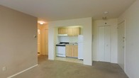 This single-floor residential unit offers a practical and functional layout designed to maximize the use of available space. The main living area is finished with wall-to-wall carpet and neutral-toned walls that lend a calm, understated ambiance throughout.

The kitchen is neatly tucked into a recessed nook, creating a defined cooking area without enclosing it entirely from the living space. It features light wood upper and lower cabinetry providing ample storage, a white gas range, a single-basin sink, and a full-size white refrigerator. The kitchen floor transitions to a smooth tile surface, distinguishing it from the carpeted living area.

A hallway visible from the main living area leads to additional rooms, suggesting a separation between living and private spaces. A flush-mount ceiling light provides overhead illumination in the living area, while the overall layout benefits from a straightforward, easy-to-navigate floor plan.

A door located on the far wall of the living area, likely a closet or utility space, adds to the storage options available within the unit. The clean, simple finishes and neutral palette throughout make this a versatile space well-suited for a variety of living arrangements.