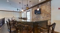 Located at 15 Brasseler Blvd in Savannah, Georgia, this single-floor space delivers a warm and inviting atmosphere through a carefully curated blend of rustic and classic design elements. The interior is anchored by a striking exposed brick accent wall that adds rich texture and a timeless character to the space.

A curved bar or counter area serves as a central focal point, finished with deep green granite countertops that complement the warm wood tones found throughout. Ornate wooden bar chairs with carved detailing reinforce the space's traditional aesthetic, adding a sense of craftsmanship and elegance.

Overhead, decorative wrought iron pendant lights hang from dark wood beams, casting a warm ambient glow that enhances the room's inviting feel. A ceiling fan with matching wrought iron detailing contributes to both comfort and visual cohesion. Recessed lighting supplements the pendant fixtures, ensuring the space is well-lit from multiple sources.

Natural light enters through windows fitted with shutters, providing a bright yet controlled atmosphere. A wall-mounted flat-screen television integrated into the brick wall adds a modern functional element without disrupting the classic design. The overall ambiance of the space is one of refined comfort, combining traditional Southern charm with practical, well-appointed features throughout.