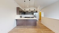 This two-story residence is thoughtfully organized across two levels, with the main living spaces occupying the upper floor and a compact entry area below. A carpeted staircase with a sleek white handrail connects the levels, leading upward into the primary living area.

The main floor features light hardwood-style flooring throughout the open living and kitchen areas. The kitchen is a standout feature, equipped with dark espresso shaker-style cabinetry, white countertops, stainless steel appliances including a range, microwave, and refrigerator, and a peninsula island with an undermount sink illuminated by a trio of pendant lights. The open layout allows the kitchen to flow naturally into the living area, which receives natural light through a window with outdoor views of greenery.

The bedroom is carpeted and comfortably sized, fitted with a ceiling fan, natural light from a window with blinds, and direct access to a dedicated closet. The full bathroom includes a tub/shower combination with chrome fixtures and wood-style flooring. A separate laundry room houses a full-size washer and dryer with overhead wire shelving for additional storage.