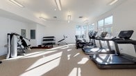 This fitness center is designed to support a comprehensive workout experience within a bright, open layout. The space is finished with clean white walls and durable speckled flooring, creating a modern and hygienic environment. Large windows line one side of the room, flooding the interior with natural light and offering views of the surrounding neighborhood, enhancing the ambiance during workouts.

The cardio section is well-stocked with multiple treadmills, elliptical trainers, and stationary bikes, all positioned to take advantage of the natural light and outdoor views. A dedicated strength training area features a cable and pulley machine system along with additional resistance equipment, providing options for a full range of targeted muscle exercises. A free weight station includes a tiered dumbbell rack with a variety of weights and an adjustable bench, accommodating users of different fitness levels.

Overhead, linear pendant lighting supplements the natural light, ensuring the space remains well-lit throughout the day. A glass-paneled entry door with direct outdoor access adds convenience and ventilation. The overall layout is spacious and uncluttered, allowing for comfortable movement between equipment stations.
