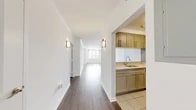 Located at 1 City Place in White Plains, NY, this one-bedroom, one-bathroom residence offers a well-planned layout across a single floor. Rich hardwood flooring flows throughout the main living areas, complemented by crisp white walls and elegant crown molding that lend the space a clean, polished character. A welcoming hallway leads into the open-concept kitchen and living room, where a pendant light fixture anchors the space above a pass-through kitchen counter. The kitchen features sage-green cabinetry, light countertops, stainless steel appliances — including a full-size refrigerator and range — and tile flooring, providing a functional and stylish cooking area. The expansive living area is bathed in natural light from large windows that frame elevated city views of White Plains, creating an airy and bright atmosphere. The bedroom is a comfortable, well-proportioned retreat, also enjoying city views through a broad window with hardwood floors continuing seamlessly. The bathroom is finished with warm beige marble-style tile, a soaking tub with shower, a classic pedestal sink, and a round mirror. Additional highlights include multiple closets offering generous storage, a dedicated laundry area, and wall sconce lighting throughout the hallways.