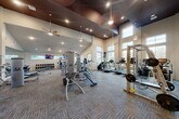 Located at 14698 Briar Forest Drive in Houston, TX, this fitness center offers a generously sized, single-floor layout designed to support a full range of workout routines. The space features soaring ceilings with a striking two-tone design — a rich dark upper section contrasting with bright white lower walls — creating a dynamic and energizing atmosphere. Pendant lights, recessed lighting, and ceiling fans are distributed throughout, ensuring the space remains well-lit and comfortable during workouts.

Large windows line multiple walls, flooding the interior with natural light and providing views of the surrounding exterior. The flooring consists of durable, patterned carpet tiles suited for a high-traffic fitness environment.

The gym is comprehensively equipped for both strength and cardiovascular training. Cybex strength machines are prominently featured alongside multi-use cable systems, adjustable benches, and a broad selection of free weights including extensive dumbbell racks reflected by floor-to-ceiling mirrors. The cardio section includes treadmills, a stair climber, and a stationary bike. Wall-mounted flat-screen televisions are positioned throughout the space for entertainment during workouts. The facility is accessed through glass-paneled doors, adding a clean, professional finish to the overall design.