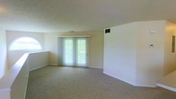 This single-floor space features a spacious, open living area finished with neutral-toned wall-to-wall carpeting that contributes to a warm, understated ambiance. The room is anchored by a set of French doors dressed with vertical blinds, allowing natural light to filter in while providing a connection to the outdoors. An arched accent window on the adjacent wall adds architectural character and introduces additional natural light. The walls are painted in a soft, neutral tone, complementing the light-colored ceiling, which features a subtle textured finish. HVAC vents are positioned both on the ceiling and wall, ensuring efficient climate control throughout the space. Clean white baseboards frame the perimeter of the room, lending a crisp, finished look. The open layout offers generous flexibility for furniture arrangement, making the space highly adaptable for a variety of uses. Overall, the space presents a clean, well-lit, and functional environment with a simple, classic aesthetic.