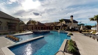 This well-appointed residential community amenity space offers a thoughtfully designed experience across its single-floor layout. At the heart of the interior is a dedicated mail center, finished with light wood-look flooring and neutral walls that create a clean, bright atmosphere. The room is lined with rows of individual mailboxes and parcel lockers, complemented by a central white countertop surface for convenient sorting. Recessed lighting and a glass-paneled door connecting to an adjacent carpeted corridor ensure the space remains functional and accessible.

Outside, the community transforms into a resort-like retreat. A sweeping, freeform swimming pool takes center stage, featuring a connected spa and flanked by lush tropical palm trees. Ample lounge seating along the pool deck invites relaxation, while a prominent outdoor fireplace adds a striking architectural focal point to the clubhouse backdrop.

A covered terrace extends the outdoor living experience along the waterfront, supported by decorative stone columns and equipped with a ceiling fan for year-round comfort. This shaded patio overlooks a serene lake with picturesque views of the surrounding neighborhood, offering both dining-height seating and cushioned chaise lounges — an ideal setting for outdoor leisure with a tranquil, scenic ambiance.
