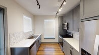 This single-story space in Denton, Texas, showcases a clean, contemporary design with consistent finishes carried throughout. The galley-style kitchen serves as a standout feature, lined on both sides with gray shaker cabinetry topped with white countertops and accented by a marble-look tile backsplash. Stainless steel appliances, including a built-in microwave, electric range, and dishwasher, provide modern functionality. Track lighting runs along the ceiling, illuminating the space efficiently, while multiple windows fitted with white horizontal blinds allow natural light to fill the room. Warm-toned wood-look flooring extends throughout the kitchen and into the living and bedroom areas, adding cohesion and visual warmth.

The bedroom is a well-proportioned, quiet space featuring the same wood-look flooring, a large window for natural light, and a ceiling fan with an integrated light fixture. The bathroom offers a tub-shower combination with full tile surround, chrome fixtures, and grab bars for added accessibility. Gray cabinetry with a white quartz-style vanity top mirrors the kitchen's design language, maintaining a unified aesthetic. Overall, the space presents a modern, functional, and well-appointed interior.