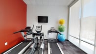 The fitness center at Forte Apartments, located at 1202 Shelby St in Indianapolis, Indiana, offers residents a dedicated space for exercise and wellness. The room features a clean, modern aesthetic with white walls accented by a bold red wall, adding an energetic visual element to the workout environment. The space is equipped with multiple cardio machines, including treadmills and an elliptical trainer, all from the Spirit brand, providing residents with quality equipment for their fitness routines. A wall-mounted flat-screen television is positioned for convenient viewing during workouts. A rack of exercise balls in bright yellow, green, and blue adds both color and versatility to the space, supporting stretching and functional training exercises. A large window along one wall allows natural light to fill the room, contributing to an open and inviting atmosphere. The flooring features a durable, dark-toned carpet designed to withstand the demands of regular exercise activity. A drop ceiling with recessed lighting ensures consistent illumination throughout. Overall, the fitness center provides a functional and well-appointed environment for residents seeking convenient on-site workout amenities.