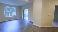 This single-floor space offers a clean, modern aesthetic defined by rich dark hardwood-style flooring that runs throughout the main living area. The walls are finished in a soft, neutral tone that creates a calm and welcoming atmosphere, complementing the crisp white baseboards and trim. Natural light flows generously into the space through a combination of a side window and a full sliding glass door, the latter providing direct access to an outdoor area while framing lush greenery beyond. The sliding door arrangement enhances the sense of openness and connectivity between the interior living space and the exterior. A ceiling fan is centrally positioned overhead, adding both functionality and a subtle design detail to the room. The open layout of the living area allows for flexible furniture arrangements and contributes to an airy, uncluttered feel. The overall ambiance is bright, fresh, and move-in ready, with thoughtful finishes that balance practicality and style throughout the space.