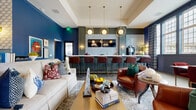 Located at 5056 Westerville Rd in Columbus, Ohio, this single-floor community clubhouse spans a generously sized footprint across five distinct areas, each exuding its own character while maintaining a cohesive, sophisticated aesthetic. The grand entrance lobby welcomes visitors with soaring ceilings, large leaded-glass windows, rich hardwood-style flooring, a brass chandelier, and bold navy blue cabinetry at the reception counter — setting an immediate tone of refined elegance. Adjacent to the entry is a stylish lounge area anchored by deep blue walls, curated gallery art, plush green velvet seating, and layered vintage-style rugs, with views into a glass-enclosed fitness center. The bar and social lounge features sage-green cabinetry, white quartz countertops, geometric gold pendant lighting, a beverage station with a commercial coffee machine and beverage refrigerator, and bar seating — ideal for community gatherings. A dedicated media room offers a large projection screen, rich crimson velvet sofas, tufted leather ottomans, and moody charcoal walls with natural light filtering through tall windows. Completing the space, the leasing office is outfitted with bold cobalt blue cabinetry, brass hardware, white countertops, and warm chandelier lighting — a polished and functional workspace that reflects the overall design excellence of this exceptional clubhouse.