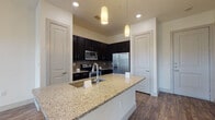 This single-floor residence offers a thoughtfully designed layout that blends modern aesthetics with everyday functionality. The heart of the home is an open-concept kitchen that immediately draws attention with its striking contrast of dark espresso cabinetry and light granite countertops. A generous island with an undermount sink serves as both a prep area and a gathering point, illuminated by elegant pendant lighting overhead. The kitchen is fully equipped with a complete suite of stainless steel appliances, including a French door refrigerator, electric range, and over-the-range microwave, all set against a tasteful tile backsplash. Wood-look flooring flows throughout the main living and kitchen areas, adding warmth and continuity to the space. The bedroom is carpeted for comfort and features a large window fitted with horizontal blinds, allowing for ample natural light and pleasant outdoor views. A hallway leads to the bathroom, which is finished with light tile flooring and dark cabinetry topped with a granite vanity, maintaining a cohesive design language. Adjacent to the bathroom, a walk-in closet provides generous storage with built-in shelving and hanging rods. Additional built-in shelving in the hallway offers practical storage solutions throughout this well-appointed residence.