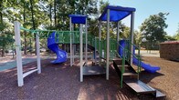 Located at 19 Landmark Dr in Columbia, South Carolina, this well-equipped outdoor recreational space offers a variety of amenities designed for active play and leisure. At the heart of the area stands a multi-level playground structure featuring blue and green accents, equipped with a spiraling blue slide, staircase access points with metal grating, elevated platforms, and blue shade canopies overhead. The play structure is surrounded by a generous wood chip safety surface that cushions the entire play zone. Nearby, a swing set with both standard and bucket seats provides additional entertainment options for younger children. A standalone climbing structure adds yet another engaging activity to the space. The playground area transitions into a hard-surface zone where a tennis court with chain-link fencing is available for recreational play. A covered picnic or shade structure with a colorful canopy provides a rest area, complemented by benches placed throughout the grounds. Mature trees line the perimeter of the space, offering natural shade and a park-like atmosphere. The surrounding landscape includes well-maintained shrubs and open green areas. The overall setting is bright, open, and inviting, making it a comprehensive outdoor recreational destination for residents of the surrounding community.