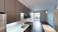 This contemporary single-floor space showcases a thoughtfully designed open layout with a strong emphasis on modern aesthetics and functional living. The kitchen is a standout feature, outfitted with rich, dark wood flat-front cabinetry that extends to the upper walls, offering generous storage. Brushed stainless steel hardware and appliances — including a gas range, built-in microwave, and wall oven — lend a polished, professional feel to the cooking area. A textured stone tile backsplash adds visual depth and contrast against the crisp white countertops, while the stainless steel undermount sink with a contemporary arc faucet completes the refined look. Dark hardwood flooring runs throughout, providing warmth and continuity across the open space. Natural light floods the interior through a large sliding glass door at the far end of the room, which opens to a private outdoor area, seamlessly blending indoor and outdoor living. The neutral gray walls and clean white ceiling keep the overall palette fresh and versatile, creating a bright, welcoming ambiance. This well-appointed space combines contemporary design elements with practical functionality throughout.