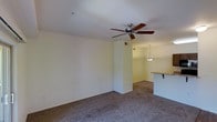 This single-floor space features an open-concept layout that connects the living area to the kitchen, creating a functional and cohesive floor plan. The living area is finished with wall-to-wall carpeting in a neutral grey tone, providing a comfortable and understated foundation for the room. A ceiling fan with wooden blades is centrally mounted, offering both practical airflow and a subtle design element overhead.

A half-wall with a bar-style counter separates the kitchen from the living space, allowing for an open sightline between the two areas while defining each zone. The kitchen area is distinguished by warm-toned walls and is illuminated by a pendant light fixture that hangs above the counter, adding a focused point of light to the space. The neutral, light-colored walls throughout contribute to a clean and airy atmosphere.

The layout is straightforward and practical, with visible HVAC vents integrated into the walls and ceiling. Overall, the space offers a simple, well-organized interior that balances openness with defined functional areas, making it adaptable for a variety of uses.