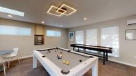Located at 555 Elmira Rd in Vacaville, California, this single-floor recreational space is thoughtfully designed for entertainment and social interaction. The main game area is anchored by a sleek white pool table with a gray felt surface, complemented by a wall-mounted cue rack. A striking geometric LED ceiling fixture serves as the room's focal point, casting warm, even light throughout. Natural light filters in through skylights and frosted roller-shade windows, enhancing the bright, open atmosphere.

Adjacent to the pool table sits a full-length shuffleboard table with a natural wood surface and dark base, positioned along a wall of diffused-light windows. A wall-mounted mini-split system ensures year-round climate comfort in this area.

A cozy lounge nook features a built-in cushioned bench, a round table with modern white chairs, and bar-height counter seating — perfect for spectators or casual conversation. Decorative wall-mounted planters and a globe pendant light add personality to this corner.

A separate media alcove is finished in a calming sage green accent wall and includes wall-mounted flat-screen televisions, an assortment of casual seating including fabric poufs and natural wood stump stools, and recessed lighting. Carpeted flooring runs throughout the space, adding warmth and acoustic comfort. A glass-paned exterior door provides convenient outdoor access.