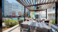 This thoughtfully designed amenity space combines stylish outdoor and interior environments to support both relaxation and productivity. The outdoor area is anchored by a substantial wood-beam pergola fitted with a ceiling fan, providing shaded comfort over a generous patio. Beneath it, a mix of dining tables with coordinating chairs and plush wicker lounge seating with colorful accent pillows creates a versatile gathering area. The patio overlooks a resort-style pool enclosed by black iron fencing, with lush landscaping and the surrounding multi-story building facades framing the view.

The interior portion of the space features a well-appointed conference room centered around a long, sleek white table with ample seating. A wall-mounted flat-screen display, a geometric linear pendant light fixture, and wood-panel accent walls contribute to the room's polished, professional character. Adjacent areas are separated by full-height glass partition walls with metal framing, maintaining visual openness while defining distinct zones. Large windows throughout the interior flood the space with natural light and offer views of the outdoor courtyard and pool. The combination of bold accent colors, patterned flooring, and modern furnishings throughout gives the space a contemporary, welcoming atmosphere.