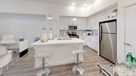 This single-floor residence is designed with a clean, modern aesthetic, combining functionality and style across its thoughtfully arranged layout. The open-concept living and kitchen area serves as the heart of the home, where light-toned hardwood-style flooring, crisp white walls, and elevated ceilings create a bright, airy atmosphere. A wall-mounted television and French doors at the far end of the living area add to its welcoming character.

The kitchen is a standout feature, outfitted with white shaker-style cabinetry extending to the ceiling, a subway tile backsplash, and stainless steel appliances including a refrigerator, range, and microwave. A large central island with a white quartz countertop, an undermount sink, and modern pendant lighting above provides generous workspace and casual seating with stylish bar stools.

The bathroom maintains the home's cohesive palette, featuring wood-look flooring, a full soaking tub with shower combo, a white vanity with ample drawer storage, and tall built-in cabinetry for additional organization.

The bedroom offers a serene retreat with plush carpeting, neutral tones, and a bright window framing views of the surrounding cityscape, allowing abundant natural light to fill the space. Overall, the residence presents a polished, contemporary living environment with consistent finishes and a well-integrated design throughout.