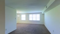 This single-floor space presents a generously sized open living area characterized by its clean, understated design. The room features neutral, light-toned walls that contribute to a bright and airy atmosphere, complemented by wall-to-wall carpeting in a warm, medium-toned gray that adds a sense of comfort underfoot. Natural light is a notable feature of this space, provided by three windows along the main wall — a single window to the left and a wider three-pane window to the right — which collectively allow substantial daylight to fill the room. A ceiling-mounted pendant light fixture with a simple bowl-shaped shade serves as a functional and modest focal point. Visible HVAC vents in the ceiling indicate a ducted climate control system. The walls are clean and unadorned, offering a blank canvas for customization. The open layout provides flexible arrangement possibilities, making it well-suited for a variety of uses. Overall, the space presents a straightforward, functional environment with a neutral aesthetic that accommodates a range of interior design preferences.
