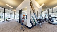 This single-floor fitness center is thoughtfully designed to support a wide range of workout routines in a bright, contemporary environment. Floor-to-ceiling steel-framed windows wrap around much of the facility, flooding the space with natural light and offering open views of the surrounding outdoor areas, creating an energizing atmosphere for exercise.

The facility is organized into distinct zones, each tailored to a specific type of training. A dedicated spin studio houses a full row of studio cycling bikes facing a wall-mounted flat-screen TV, ideal for group or self-guided cycling sessions. An adjacent cardio area features multiple treadmills and elliptical machines positioned directly in front of expansive windows, allowing users to enjoy outdoor views while training.

The strength training section is well-appointed with a rack of dumbbells spanning a wide weight range, a cable machine, a power rack with adjustable benches, and a selection of medicine balls and stability balls for functional fitness. A bold geometric graphic accent wall in tones of white, charcoal, and teal adds a dynamic visual element to the free weights area.

Throughout the space, recessed and pendant lighting complement the abundant natural light. The neutral-toned carpet flooring, clean white walls, and consistent black-framed architectural details lend the facility a polished, modern aesthetic suited for an active lifestyle.
