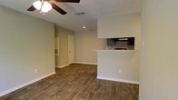 This single-story space showcases an open-concept design that seamlessly connects the living area to the kitchen. The living room features durable wood-look plank flooring that runs throughout, complemented by neutral gray-toned walls and white baseboards that create a clean, modern aesthetic. A ceiling fan with a light fixture is centrally mounted, providing both airflow and ambient lighting, while recessed lighting adds additional illumination throughout the space.

A half-wall partition separates the kitchen from the living area, topped with a light-colored granite or stone countertop that functions as a breakfast bar or pass-through. The kitchen is partially visible beyond the partition, featuring dark cabinetry and an under-cabinet range hood above the cooktop. A HVAC vent and return are conveniently positioned in the living area, ensuring efficient climate control.

A doorway visible toward the rear of the space leads to additional rooms, suggesting a functional layout with clear traffic flow. The textured ceiling adds a traditional finish to the interior. Overall, the space presents a well-maintained, move-in-ready environment with contemporary finishes throughout.