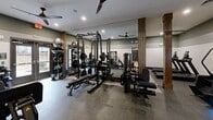 Located at 13100 Margie Ln in Raleigh, NC, this fitness center is a compact yet thoughtfully designed workout facility that offers a comprehensive range of exercise options. The space features a modern two-toned wall treatment, with a neutral white lower half and a deep sage green upper half, complemented by dark baseboards that lend a polished, contemporary aesthetic throughout.

The flooring consists of durable speckled rubber gym flooring, ideal for both heavy equipment and high-impact workouts. Recessed lighting and ceiling-mounted fans ensure the space remains well-lit and well-ventilated during use.

The cardio zone is equipped with multiple treadmills, a stair climber, and a stationary cycle, providing a variety of cardiovascular training options. The strength training area features a multi-functional cable machine, a leg press, a full power rack with bench press, and a free weight station stocked with dumbbells and medicine balls. Warm-toned wooden columns add an architectural accent that softens the otherwise utilitarian feel of the space.

Full-length mirrors line one wall of the strength training section, visually expanding the space and allowing users to monitor their form. A glass-paneled door provides access to the facility, with views suggesting proximity to outdoor amenities. The overall ambiance is clean, functional, and modern, making this fitness center a well-rounded resource for residents.