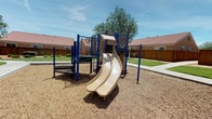 This inviting outdoor playground area is thoughtfully designed to provide a safe and engaging environment for children and families. The central play structure features a dual-lane slide and climbing platforms supported by sturdy blue metal posts, set within a wood chip safety surface bordered by a concrete curb. A second play structure with a spiral slide is positioned nearby, offering additional play options across the space.

Surrounding the play areas, well-maintained green lawns stretch outward, complemented by mature trees that provide natural shade throughout the space. A smooth concrete walking path winds gracefully through the grounds, connecting the different areas and offering easy accessibility. Interactive play elements, including a musical horn station, are integrated into the playground to encourage creative and sensory play.

The space is enclosed by wooden privacy fencing, with neighboring buildings featuring distinctive red-tiled rooftops visible in the background, contributing to a cohesive residential character. The open sky above and the generous landscaping create a bright, airy atmosphere. Overall, this playground combines functional recreational equipment with attractive green spaces, making it an appealing and practical amenity for the surrounding community.