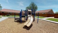 This inviting outdoor playground area is thoughtfully designed to provide a safe and engaging environment for children and families. The central play structure features a dual-lane slide and climbing platforms supported by sturdy blue metal posts, set within a wood chip safety surface bordered by a concrete curb. A second play structure with a spiral slide is positioned nearby, offering additional play options across the space.

Surrounding the play areas, well-maintained green lawns stretch outward, complemented by mature trees that provide natural shade throughout the space. A smooth concrete walking path winds gracefully through the grounds, connecting the different areas and offering easy accessibility. Interactive play elements, including a musical horn station, are integrated into the playground to encourage creative and sensory play.

The space is enclosed by wooden privacy fencing, with neighboring buildings featuring distinctive red-tiled rooftops visible in the background, contributing to a cohesive residential character. The open sky above and the generous landscaping create a bright, airy atmosphere. Overall, this playground combines functional recreational equipment with attractive green spaces, making it an appealing and practical amenity for the surrounding community.