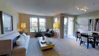 This single-floor space offers a welcoming and well-lit environment, thoughtfully designed to maximize comfort and functionality. The living area serves as the centerpiece of the space, featuring a generous, open layout with plush carpeted flooring that adds warmth underfoot. Large, multi-pane windows line the exterior wall, allowing abundant natural light to flood the interior and frame views of lush greenery outside — creating a seamless connection between indoors and the surrounding landscape. A sliding glass door provides direct access to a private balcony, further enhancing the sense of openness and offering an additional outdoor retreat. The neutral, warm-toned walls create a calm and versatile backdrop, complementing a range of furnishing styles. The living area accommodates a full seating arrangement, with ample space for a sofa, accent chair, and coffee table, making it well-suited for both relaxation and entertaining. Tall floor lamps and indoor plants add character and a touch of nature to the interior. The overall ambiance is airy, serene, and inviting, with a clean modern aesthetic that balances practicality with a comfortable residential feel.