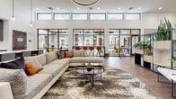 Located in Wichita, KS, this single-floor amenity clubhouse combines contemporary design with warm, inviting materials to create a sophisticated communal space. The grand entrance welcomes visitors with rich hardwood flooring, a striking solid wood door, and lush tropical greenery that sets the tone for the interiors beyond.

The main lounge area is a standout feature, boasting soaring ceilings lined with expansive clerestory windows that flood the space with natural light. A large sectional sofa, patterned area rug, and a sleek glass coffee table anchor the space, with views out to an outdoor courtyard and surrounding community. An adjoining seating area features a dramatic floor-to-ceiling stone and metal-clad fireplace with an integrated flat-screen TV, complemented by leather club chairs and a cozy coffee table arrangement.

A spacious kitchen and refreshment area offers dark cabinetry, stainless steel appliances including a full-size refrigerator, a white quartz island, and hardwood floors, making it well-equipped for entertaining. Decorative wood-textured wall art adds an artistic touch to the space.

Large concrete-tiled planters filled with dramatic foliage divide open areas, adding a natural, architectural element. Tufted leather bench seating provides additional lounging options throughout. The overall ambiance blends modern industrial design with natural warmth, creating a functional and visually compelling communal environment.