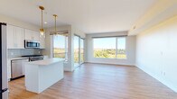 Located along Chief Justice Cushing Highway in Scituate, MA, this single-floor residence offers a thoughtfully designed open-concept layout with contemporary finishes throughout. Light hardwood flooring runs seamlessly across the living, dining, and bedroom areas, enhancing the bright, airy atmosphere created by an abundance of large windows.

The open living and dining area is a standout feature, with expansive floor-to-ceiling windows and sliding glass doors opening to a private balcony, offering sweeping panoramic views of the lush surrounding landscape. Gold pendant lighting accents the space with a warm, modern touch.

The kitchen is outfitted with white shaker-style cabinetry, light quartz countertops, a soft blue subway tile backsplash, and stainless steel appliances including a gas range, microwave, and dishwasher. A central island provides additional prep and dining space.

Multiple bedrooms feature large windows framing scenic outdoor views and light hardwood flooring. One bedroom includes an en-suite bathroom and walk-in closet, while another offers direct access to an additional bathroom. The bathrooms are finished with white tile, quartz vanity tops, frameless glass shower enclosures, and chrome fixtures, delivering a clean, modern aesthetic throughout.