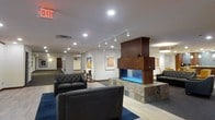 This single-floor space presents a well-appointed lounge or lobby area that blends modern design with warm, inviting elements. The centerpiece of the room is a striking freestanding fireplace, framed in rich dark wood and set on a natural stone base with a vivid blue-lit firebox, creating a bold focal point that anchors the seating area. Surrounding the fireplace, tufted black leather sofas and chairs are arranged on a deep blue area rug layered over light hardwood-style flooring, offering a comfortable gathering space.

The walls serve as a gallery, displaying framed artwork that adds color and visual interest to the otherwise neutral white surfaces. Track lighting runs along the ceiling, providing adjustable, directional illumination that highlights both the artwork and the overall space. Recessed lighting complements the track fixtures, ensuring the room is evenly and warmly lit throughout.

A corridor extending from the lounge hints at additional rooms or offices beyond, suggesting this space functions as a shared or communal area within a larger facility. Decorative sculptural accents are placed thoughtfully around the room, enhancing the curated, polished atmosphere.