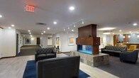 This single-floor space presents a well-appointed lounge or lobby area that blends modern design with warm, inviting elements. The centerpiece of the room is a striking freestanding fireplace, framed in rich dark wood and set on a natural stone base with a vivid blue-lit firebox, creating a bold focal point that anchors the seating area. Surrounding the fireplace, tufted black leather sofas and chairs are arranged on a deep blue area rug layered over light hardwood-style flooring, offering a comfortable gathering space.

The walls serve as a gallery, displaying framed artwork that adds color and visual interest to the otherwise neutral white surfaces. Track lighting runs along the ceiling, providing adjustable, directional illumination that highlights both the artwork and the overall space. Recessed lighting complements the track fixtures, ensuring the room is evenly and warmly lit throughout.

A corridor extending from the lounge hints at additional rooms or offices beyond, suggesting this space functions as a shared or communal area within a larger facility. Decorative sculptural accents are placed thoughtfully around the room, enhancing the curated, polished atmosphere.