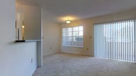 This single-floor residential space features a straightforward, functional layout centered around a spacious living area. The room is finished with wall-to-wall carpet in a neutral beige tone, contributing to a warm and understated aesthetic throughout. Smooth, light-colored walls are complemented by a textured ceiling, and a flush-mount ceiling light fixture provides overhead illumination.

Natural light is a key feature of the living area, brought in through a large window fitted with white vertical blinds, as well as an adjacent sliding glass door, also dressed in matching vertical blinds. This combination suggests access to an outdoor area such as a patio or balcony, enhancing the sense of openness. The wall layout includes a partial dividing wall or half-partition visible on the left side of the space, which may indicate a transition to a kitchen or dining area.

Electrical outlets and light switch plates are evenly distributed along the walls, reflecting a practical design. The overall ambiance of the space is clean, neutral, and move-in ready, offering a blank canvas suitable for a variety of interior arrangements.