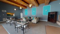 Located at 4427 Wilmington Pike in Dayton, Ohio, this single-floor community space makes an immediate impression with its striking design and bold color palette. The main gathering area is defined by soaring vaulted ceilings clad in warm natural wood planks, supported by exposed beams and cooled by multiple ceiling fans — a combination that lends the space an open, airy character. The walls are painted in a cool slate gray, enlivened by eye-catching teal accent panels framed in white, creating a lively and cohesive aesthetic throughout. Comfortable seating arrangements feature plush tan sofas accented with teal throw pillows, accompanied by a dining area with ornate wrought-iron tables and chairs. A brick accent wall near the entertainment area adds a rustic contrast to the contemporary color scheme. The kitchen is fully equipped with wood cabinetry, a refrigerator, range, and microwave, with a pass-through counter connecting it to the main gathering space. A secondary entrance area offers access to an outdoor patio, with vertical blinds and glass doors that allow natural light to filter in. The overall ambiance is welcoming, energetic, and well-suited for gatherings and community events.