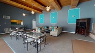 Located at 4427 Wilmington Pike in Dayton, Ohio, this single-floor community space makes an immediate impression with its striking design and bold color palette. The main gathering area is defined by soaring vaulted ceilings clad in warm natural wood planks, supported by exposed beams and cooled by multiple ceiling fans — a combination that lends the space an open, airy character. The walls are painted in a cool slate gray, enlivened by eye-catching teal accent panels framed in white, creating a lively and cohesive aesthetic throughout. Comfortable seating arrangements feature plush tan sofas accented with teal throw pillows, accompanied by a dining area with ornate wrought-iron tables and chairs. A brick accent wall near the entertainment area adds a rustic contrast to the contemporary color scheme. The kitchen is fully equipped with wood cabinetry, a refrigerator, range, and microwave, with a pass-through counter connecting it to the main gathering space. A secondary entrance area offers access to an outdoor patio, with vertical blinds and glass doors that allow natural light to filter in. The overall ambiance is welcoming, energetic, and well-suited for gatherings and community events.