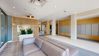 Located at 25 Woodridge Cres in Ottawa, Ontario, this well-appointed residential lobby and common area presents a thoughtfully designed single-floor layout that balances functionality with modern aesthetics. The space is defined by a clean, contemporary palette of warm golden wall panels, crisp white ceilings with recessed lighting, and a combination of large-format porcelain tile and plush carpet underfoot.

The entrance hallway is bright and welcoming, benefiting from floor-to-ceiling glazed doors that draw in natural light and offer views of the landscaped exterior. A prominent mailroom area is integrated into the hallway, featuring extensive built-in mailboxes set against the warm-toned accent walls.

The living room lounge area is a standout feature, furnished with generous, contemporary seating and anchored by a striking vertical slatted wood partition screen that adds warmth and visual texture. Lush indoor planters introduce greenery throughout, enhancing the inviting atmosphere. A wall-mounted digital display screen serves as an informational focal point within the space.

Large sliding glass windows in the lounge area provide direct sightlines into an adjacent indoor pool facility, creating a dynamic connection between the social and recreational areas. Pendant and recessed lighting throughout contribute to a well-lit, polished ambiance suited to a busy residential community.