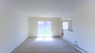 This single-floor space offers a clean, open layout designed for comfortable everyday living. The main living area is finished with wall-to-wall carpeting and crisp white walls that lend a bright, airy feel to the interior. A large sliding glass door serves as a focal point of the living room, filling the space with natural light and providing direct access to an outdoor balcony with views of a grassy, tree-lined exterior. A baseboard heating unit runs beneath the sliding door, ensuring year-round comfort. Adjacent to the living area, a pass-through opening connects to the kitchen, allowing for an open, communicative flow between spaces. The kitchen features a countertop bar ledge visible through the pass-through, along with its own window that draws in additional natural light. The overall ambiance is clean, neutral, and move-in ready, with a simple and functional design that offers a welcoming sense of openness. The white ceilings and walls reflect light effectively, making the interior feel spacious and well-illuminated throughout the day.