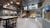 This thoughtfully converted historic Milwaukee building masterfully blends industrial heritage with modern amenities across its single floor. The centerpiece is a dramatic communal lounge boasting soaring ceilings, exposed structural columns, and original cream city brick walls. Oversized windows flood the space with natural light while offering views of the surrounding historic streetscape. Bold black pendant lighting with gold interiors hangs above a mix of high-top bar seating, casual lounge groupings, and intimate two-person tables, creating flexible zones for socializing or relaxing. Polished concrete floors and exposed ductwork reinforce the industrial aesthetic throughout. A well-equipped kitchenette features warm wood cabinetry, a commercial coffee station, open shelving, and a stainless steel sink set against the original brick backdrop. The fitness facilities include a dedicated weight room with rack systems, mirrors, and cardio machines set beneath a rustic wood-plank ceiling, as well as a spacious group fitness studio with original timber columns, rubber flooring, a large display screen, and mirrored walls. A utility support room with whitewashed stone walls rounds out the functional infrastructure of the space. Milwaukee-themed artwork throughout adds a strong sense of local identity.